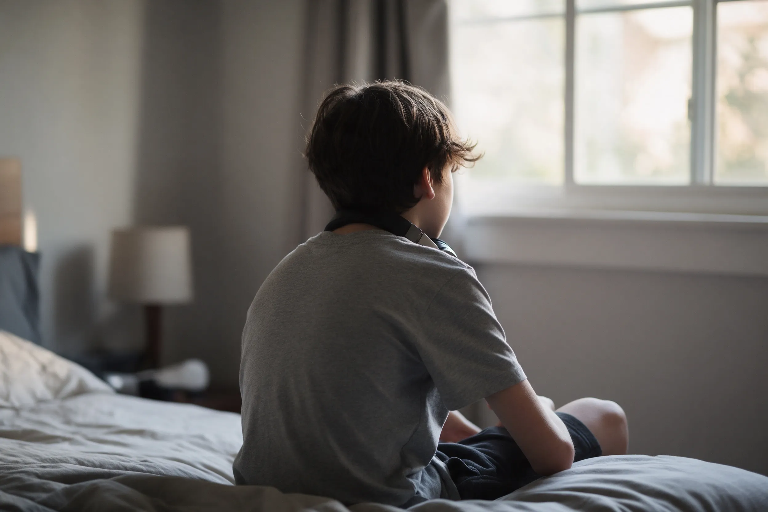 Un adolescente sentado en el borde de la cama, de espaldas, mira hacia la ventana de su habitación.