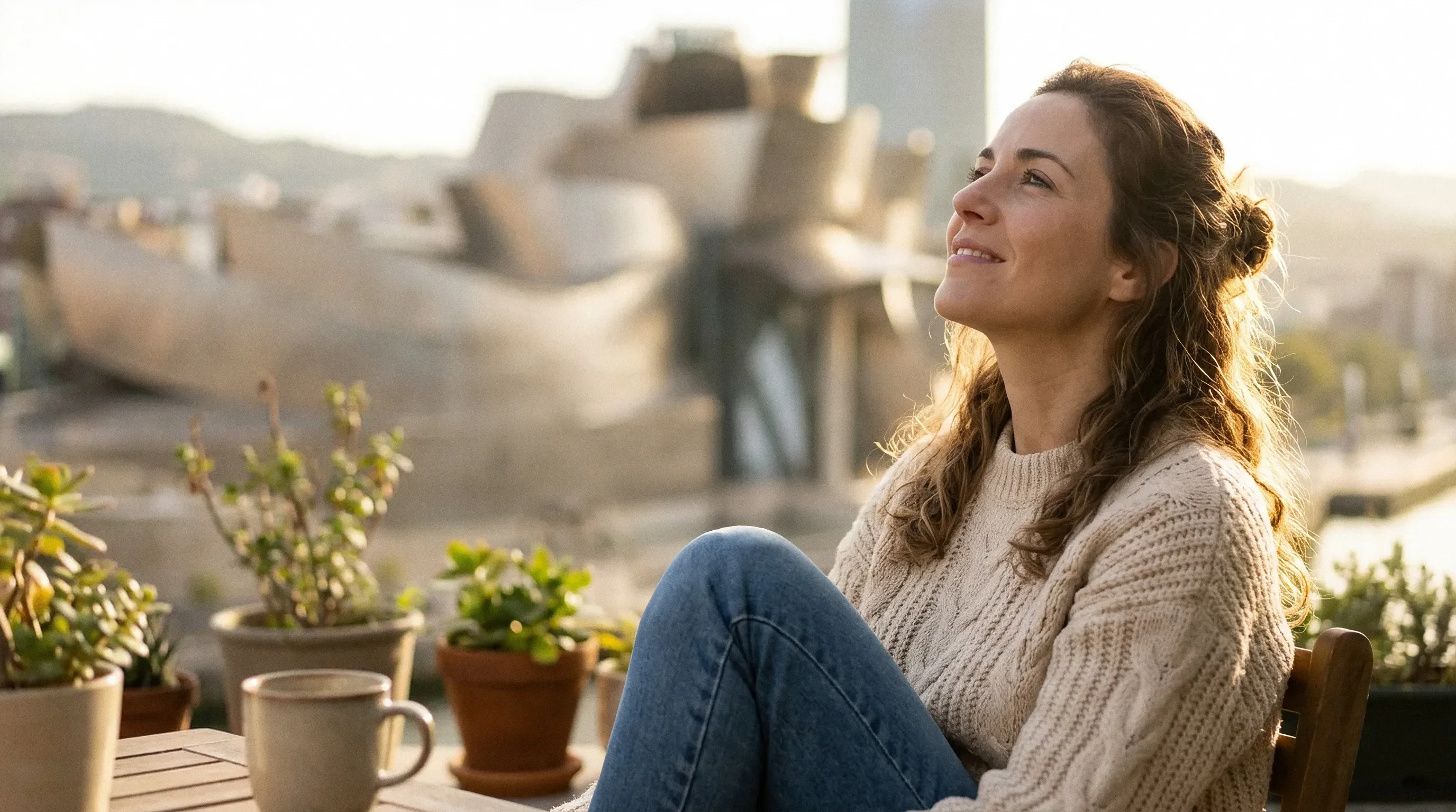 Una mujer descansa en una terraza con plantas, con el Museo Guggenheim de Bilbao al fondo en un atardecer cálido