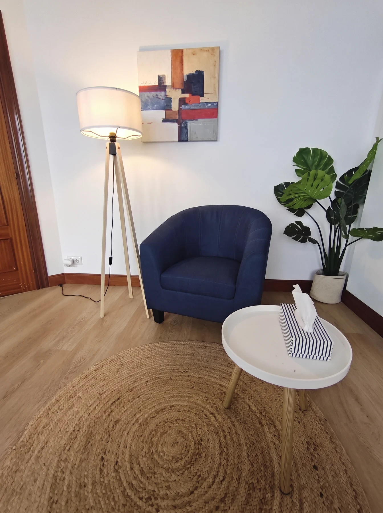 Rincón de una consulta con una butaca azul sobre una alfombra de yute, una mesita, una lámpara de pie y una planta.