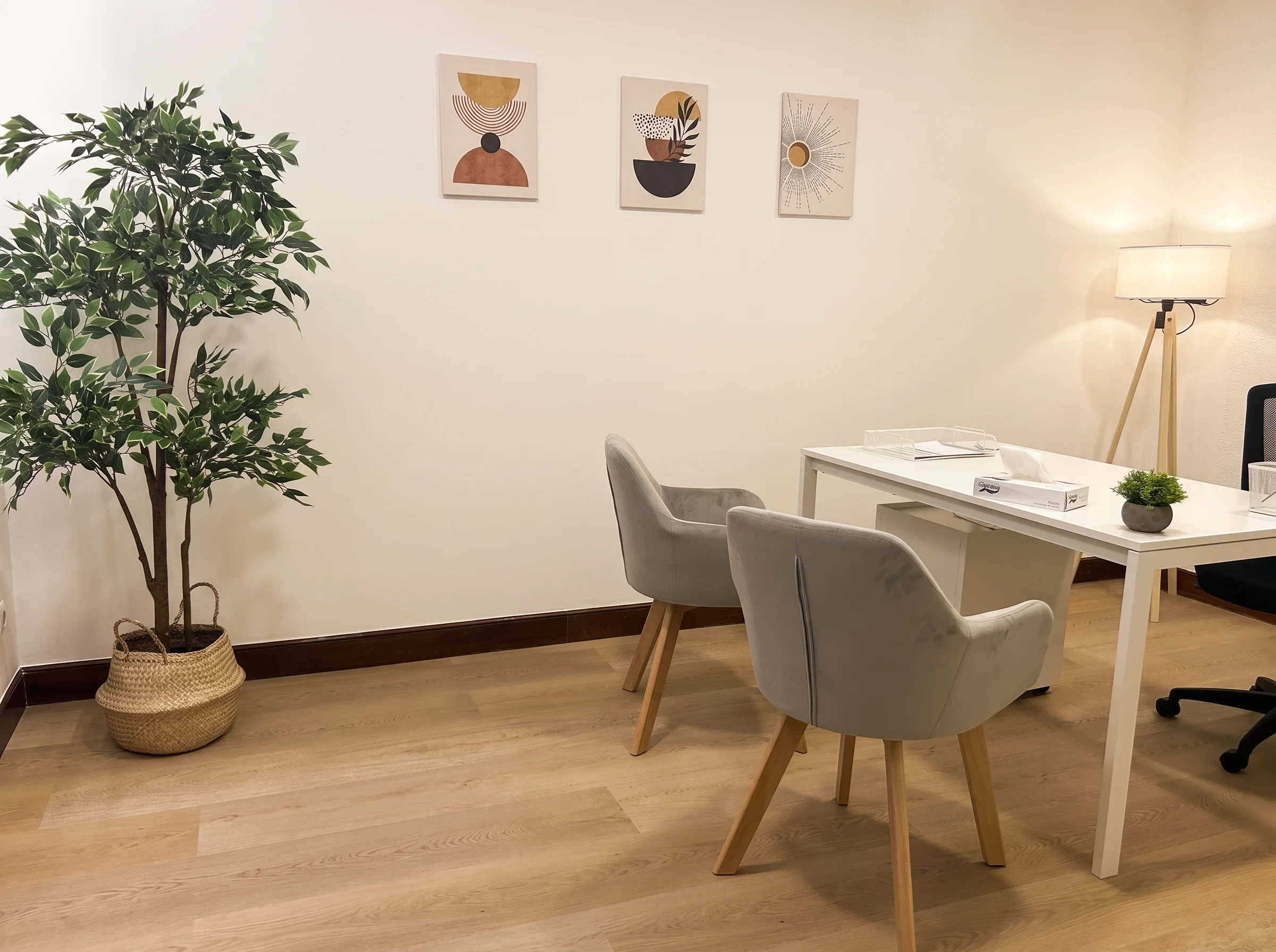 Consulta con mesa blanca, dos sillas grises, tres cuadros en tonos ocres, una lámpara de pie y una planta.