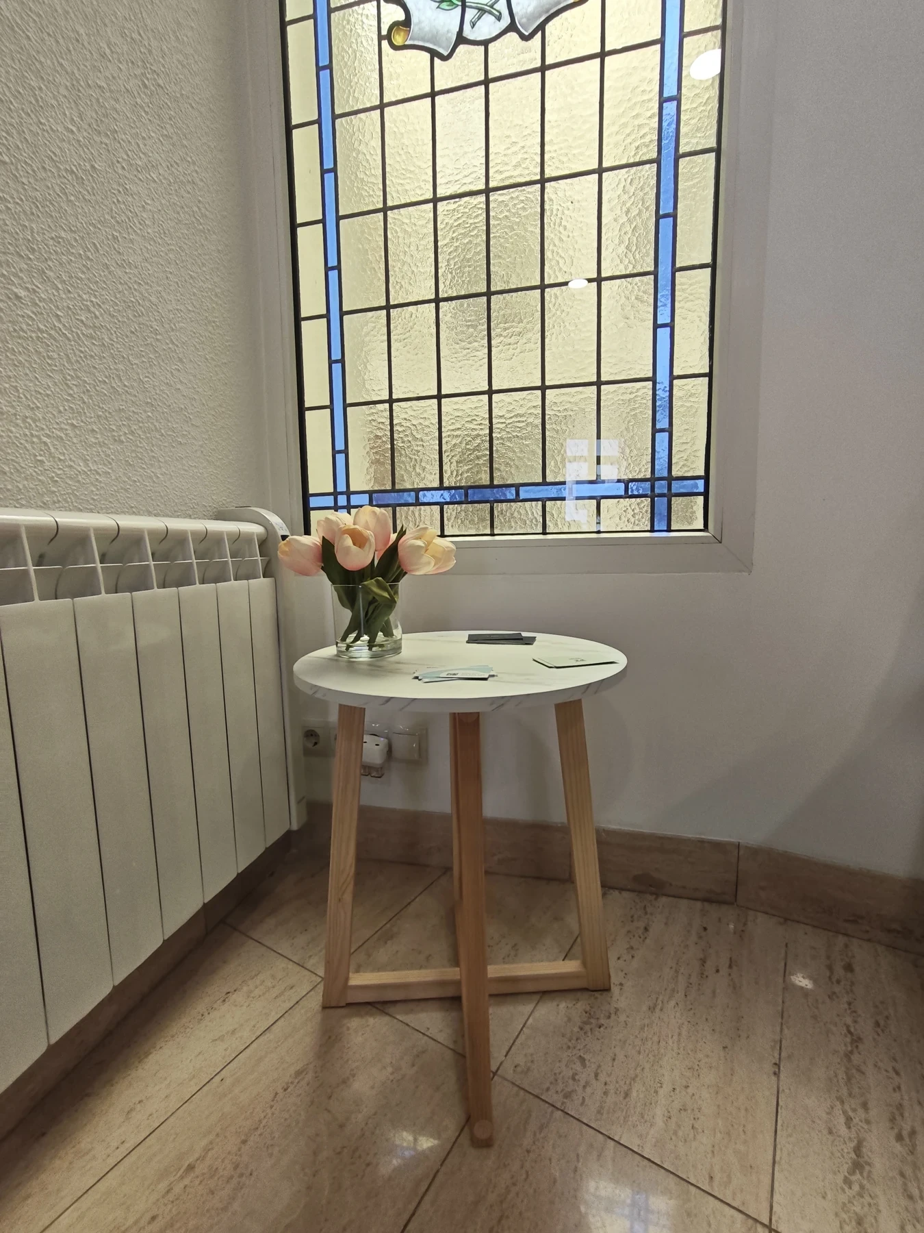 Rincón de la sala de espera con una vidriera, una mesita de madera y un jarrón con tulipanes.
