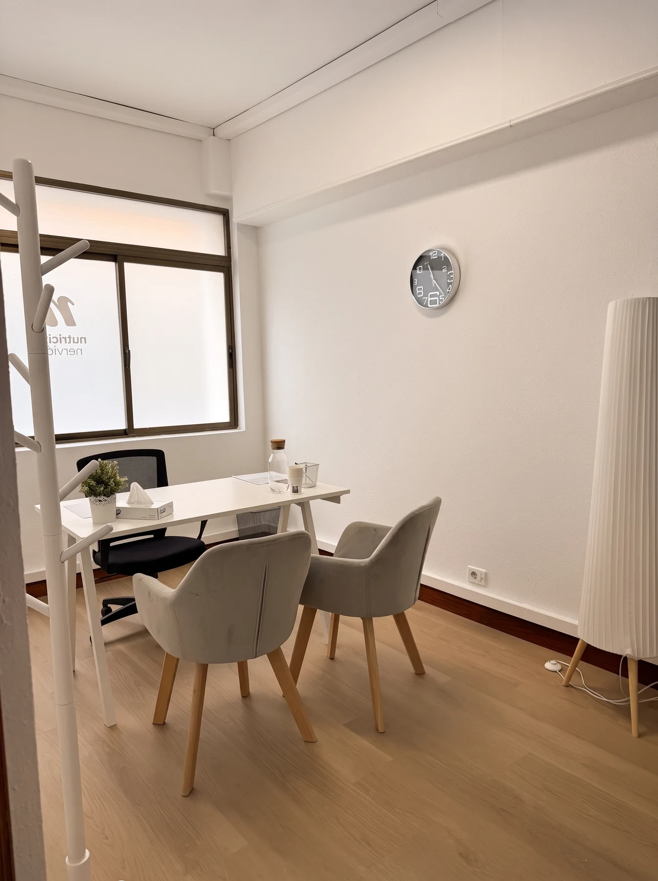 Consulta con ventana, mesa blanca, dos butacas grises, reloj de pared y lámpara de pie.