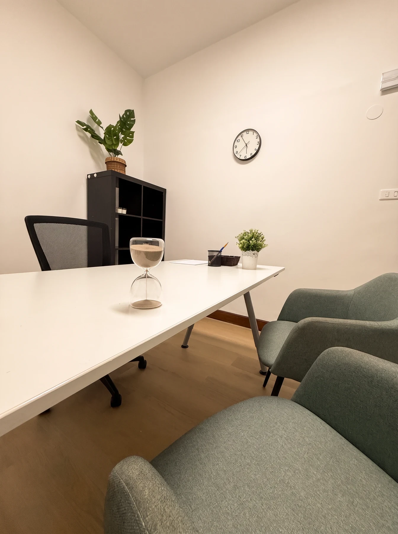 Consulta con mesa blanca, un reloj de arena, una planta y butacas verdes.