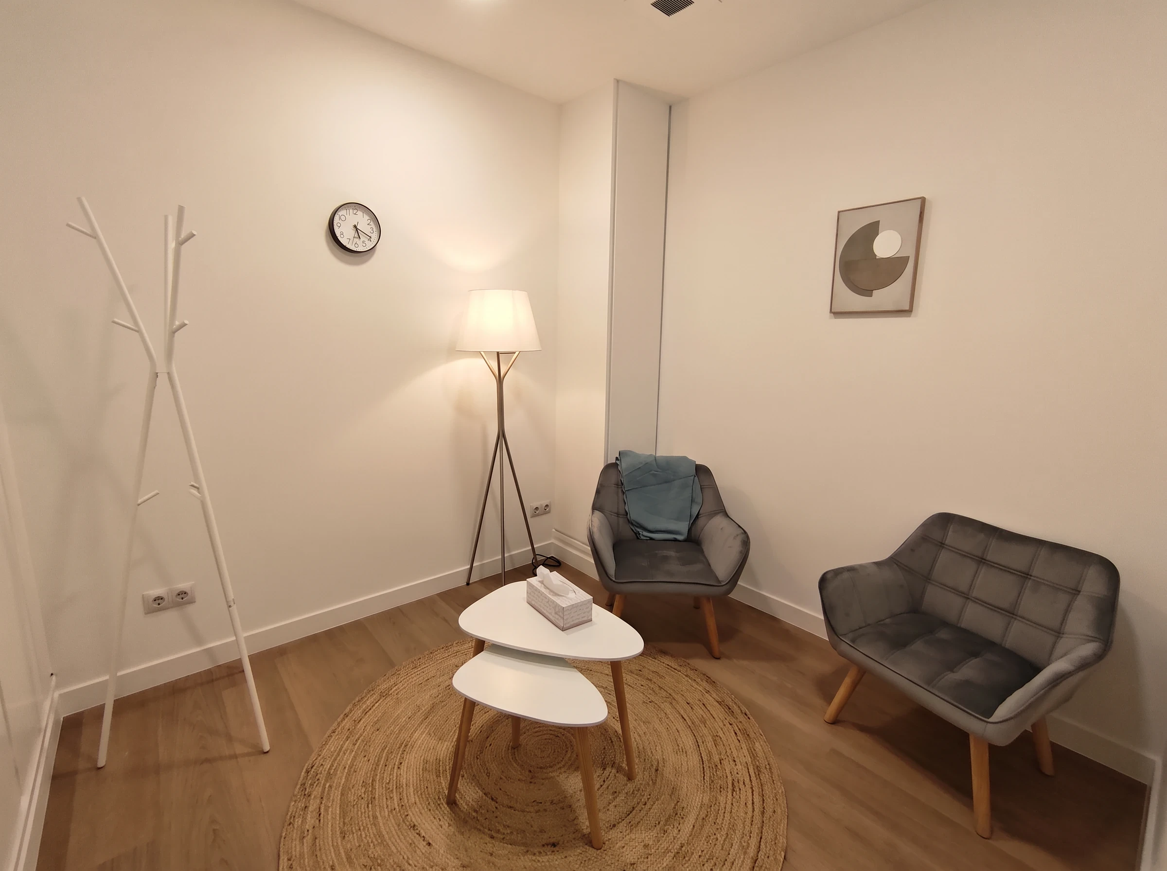Sala con dos butacas grises, una lámpara de pie encendida, mesitas blancas y una alfombra de yute.