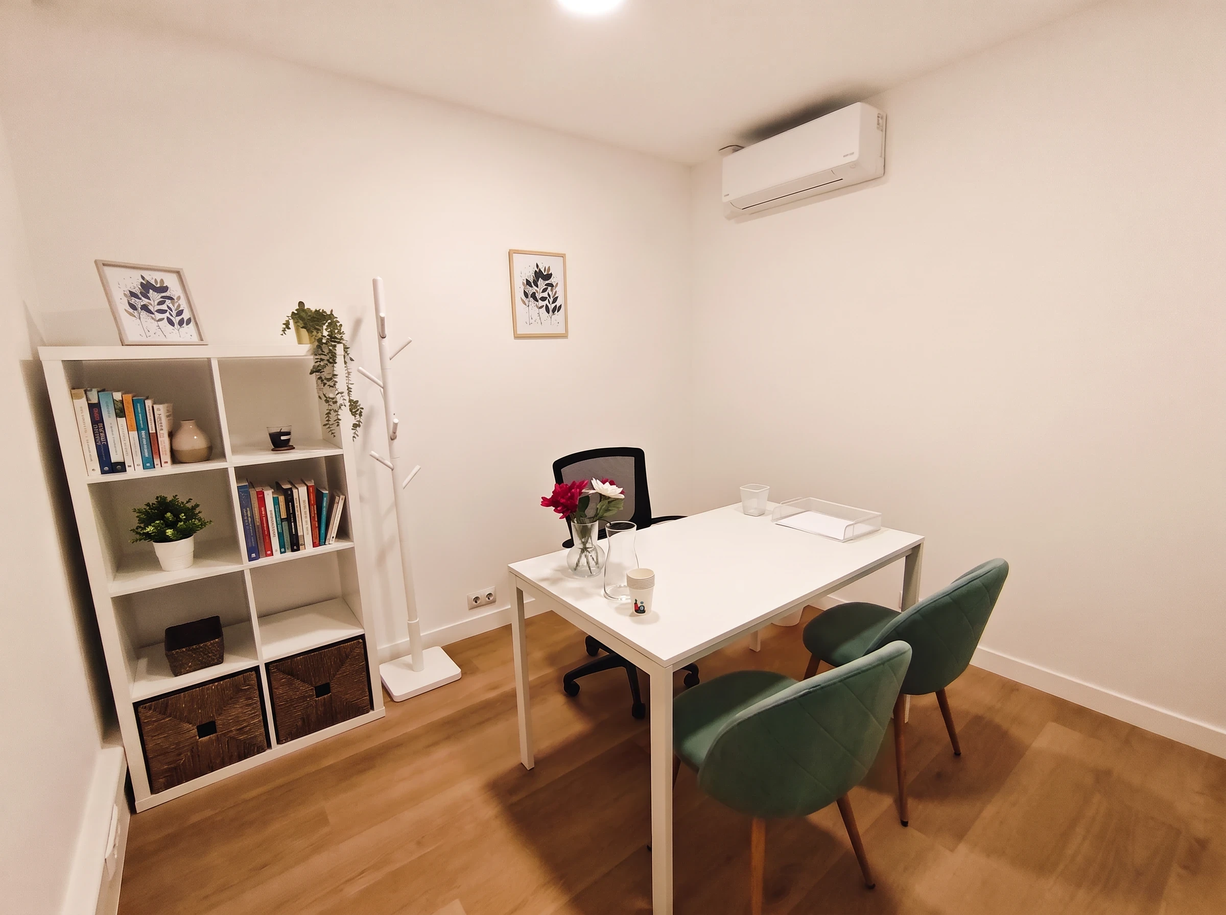 Consulta con mesa blanca, flores, dos butacas verdes y una estantería con libros, una planta y cestas.