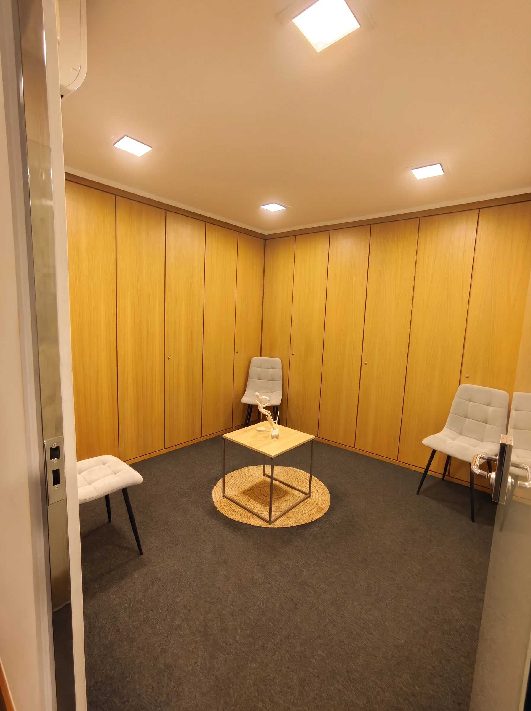 Sala pequeña con paredes de madera, sillas blancas y una mesita sobre alfombra redonda.