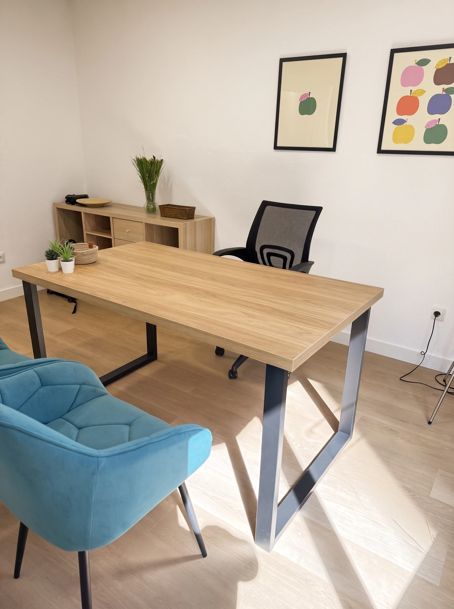 Mesa de madera con una butaca verde azulado, una silla de trabajo y cuadros de manzanas.