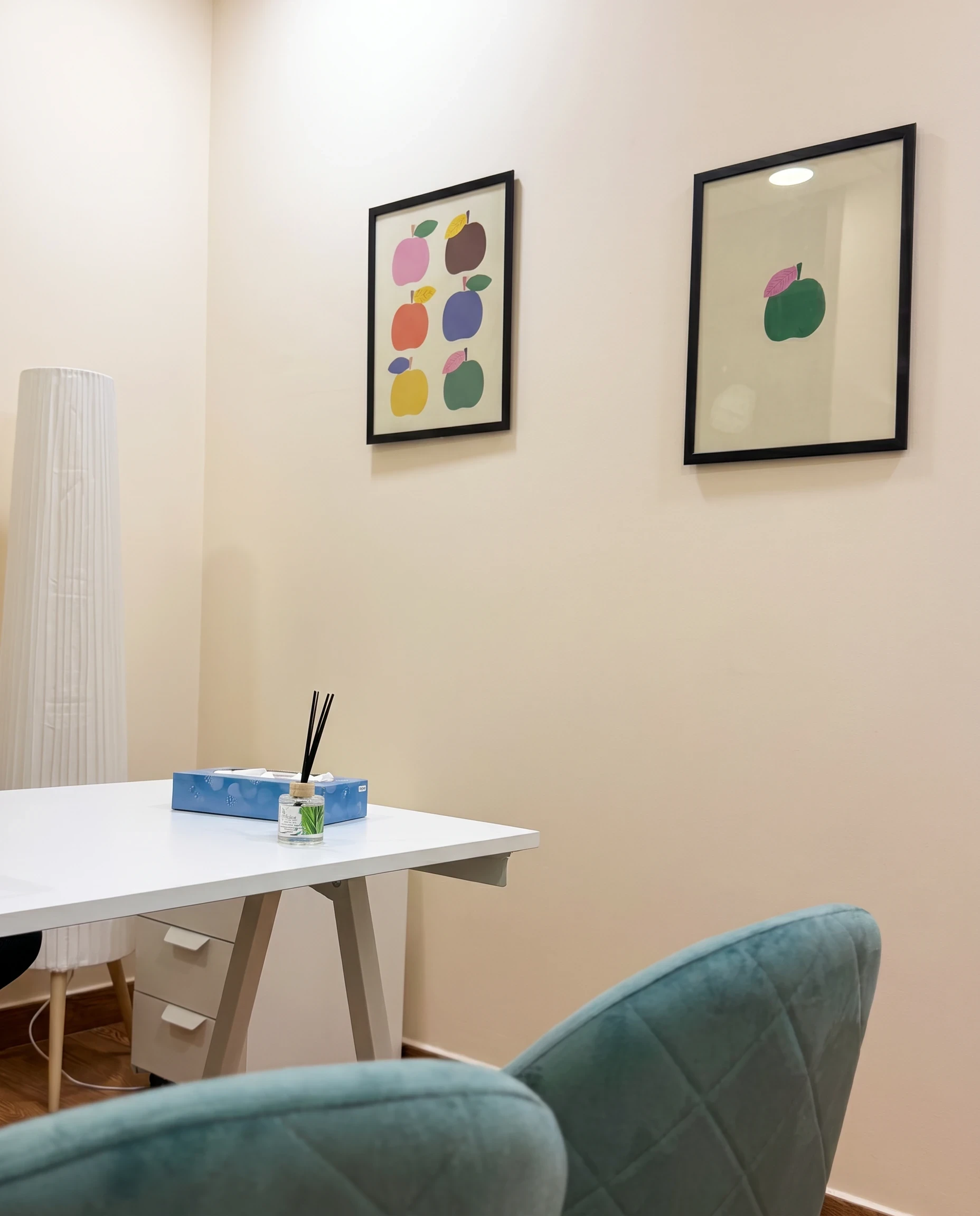 Dos cuadros con manzanas de colores sobre la pared de una consulta, junto al escritorio y las butacas turquesa.