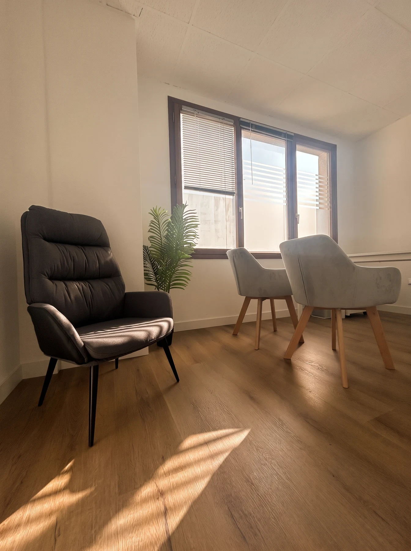 Sillón gris de respaldo alto y dos butacas junto a la ventana, con la luz del sol sobre el suelo de madera.