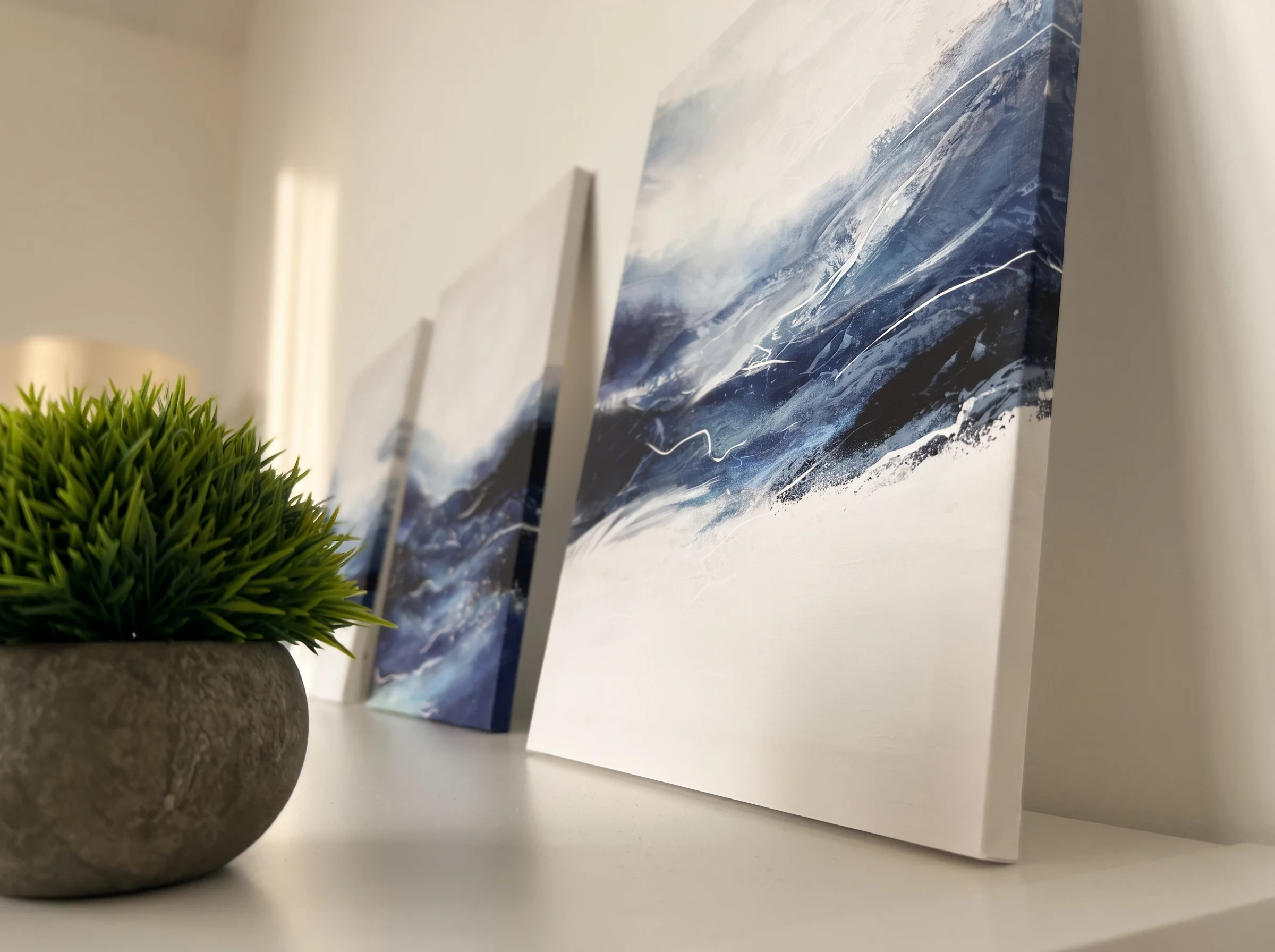 Tres lienzos abstractos en azul y blanco sobre una repisa, con una planta en primer plano.