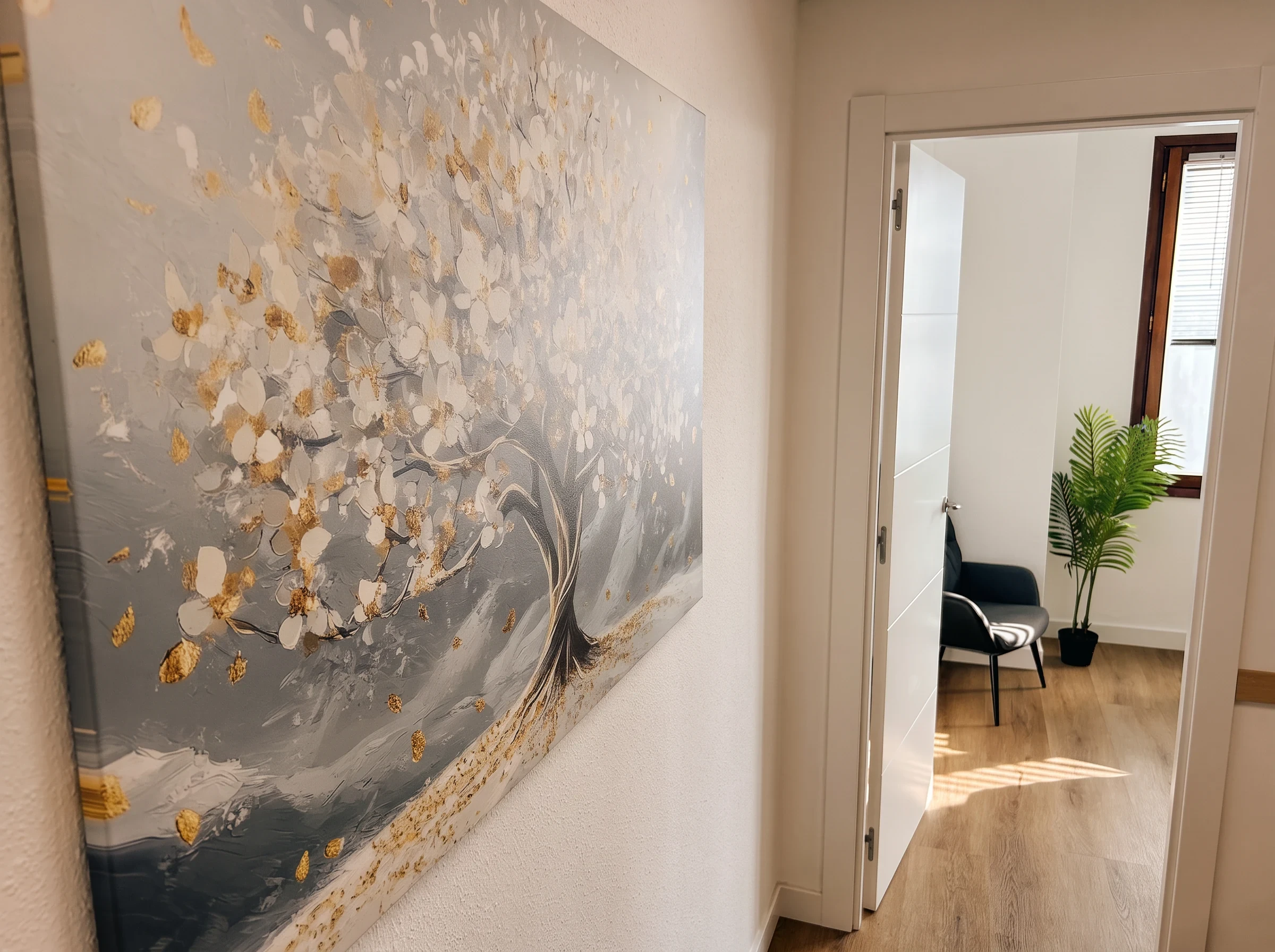 Pasillo con un cuadro de un árbol en flor y la puerta abierta a una consulta con sillón y palmera.