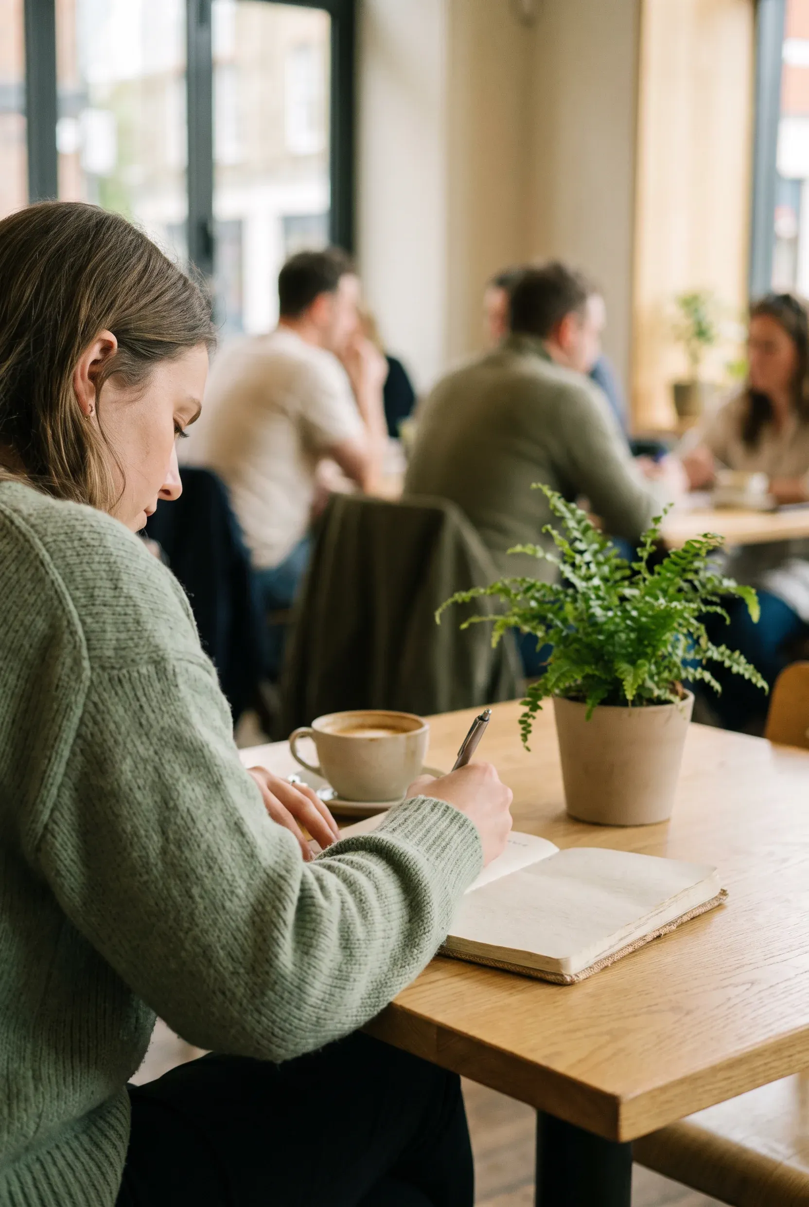 Una persona escribe en un cuaderno en la mesa de una cafetería luminosa, con una taza de café.